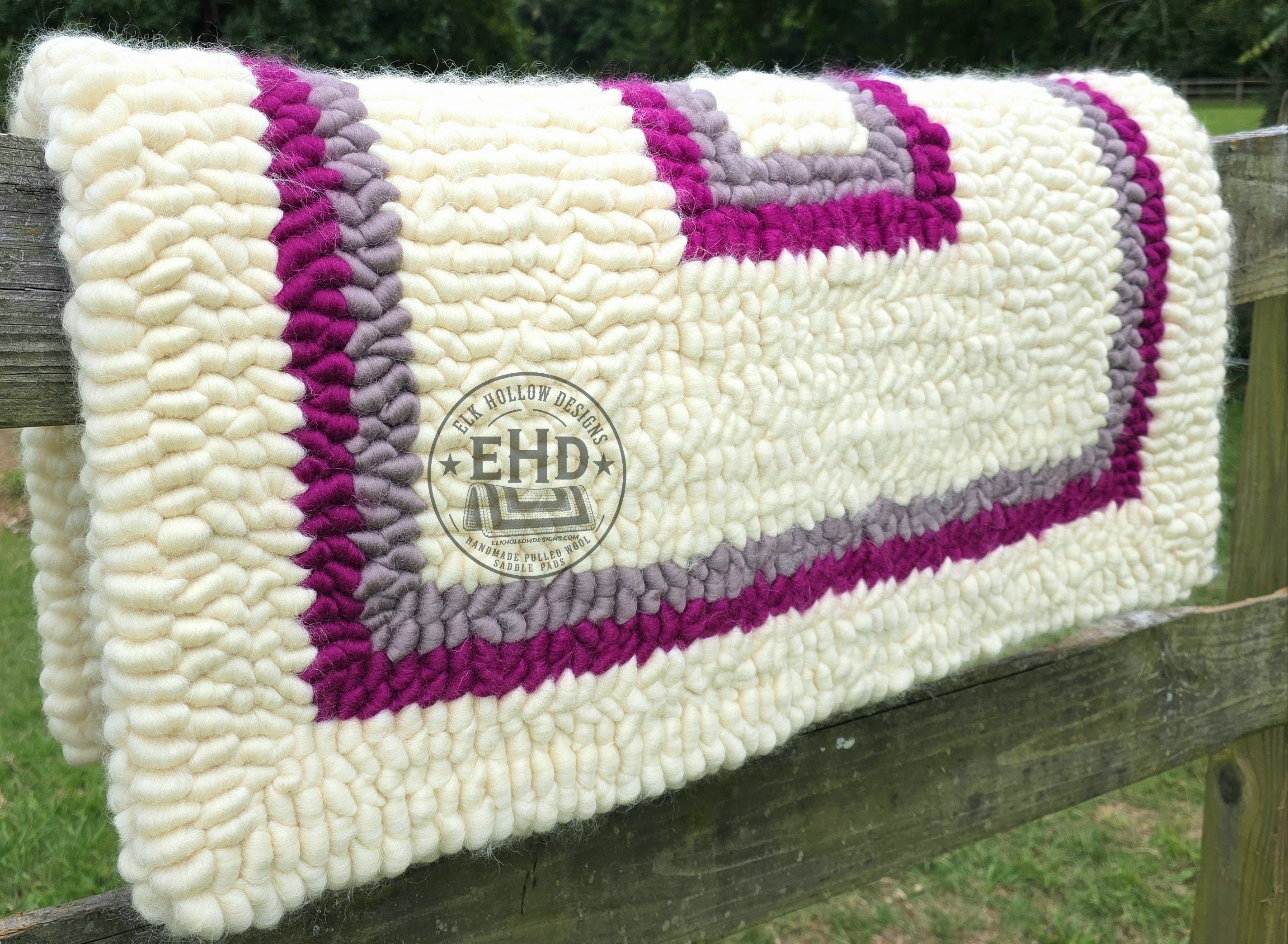 A pulled wool saddle pad with natural, magenta, and gray borders draped over a wooden fence in an outdoor setting with trees in the background.